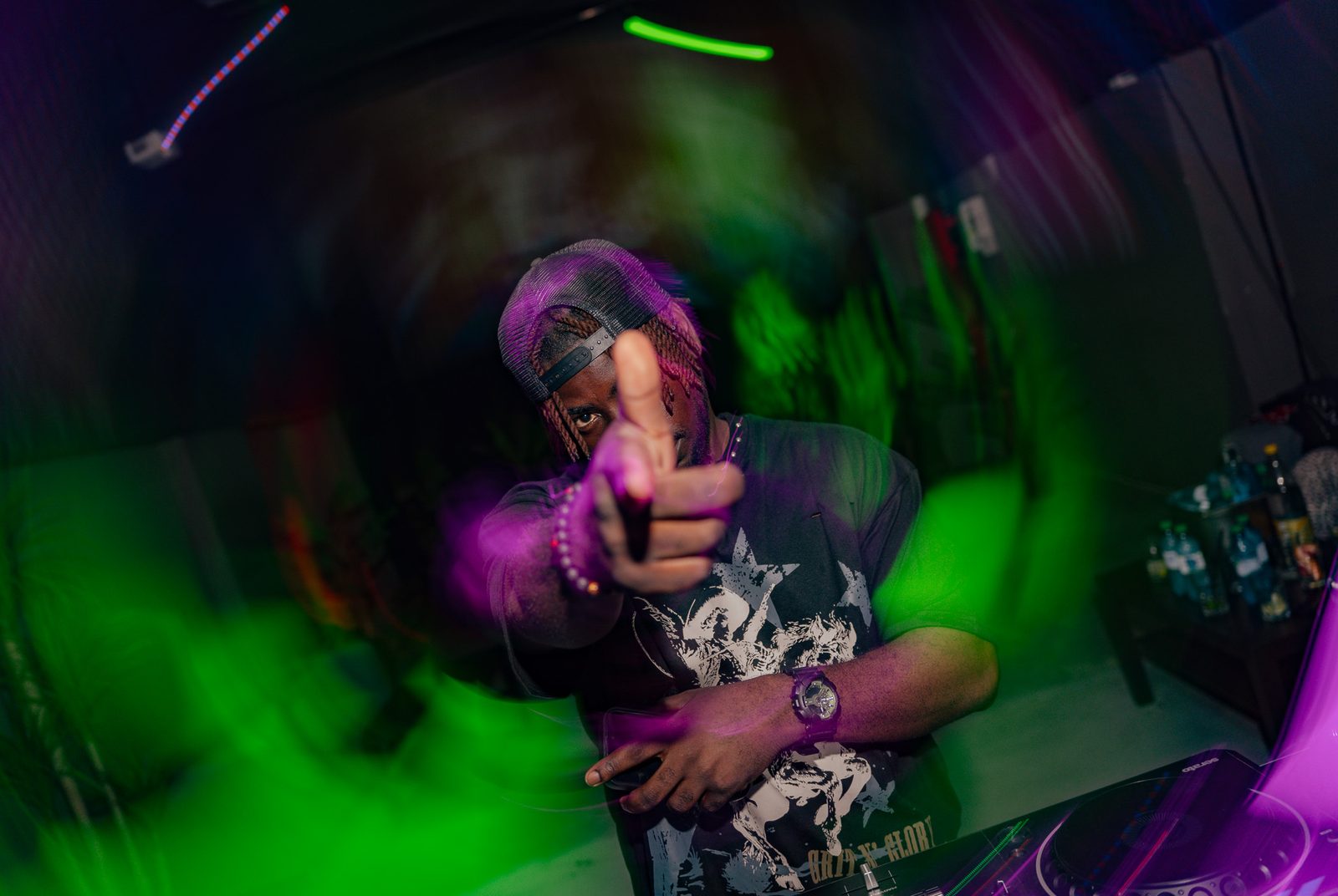 DJ Papi pointing at the camera mid-set, lit in green and purple club light